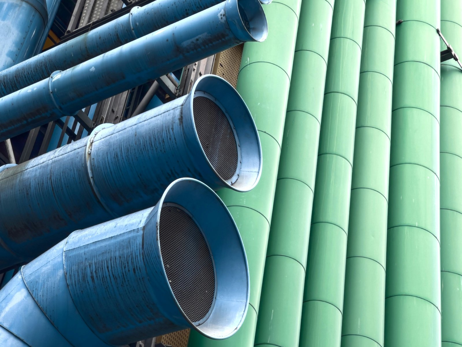 Source: Artem Horovenko (via Unsplash). On the left side of the image, there are two adjacent pairs of large blue tubes, aligned vertically, and running from the left to the center of the image, featuring a convex-fairing, and a grate over their respective openings. In the background we can see more large blue tubes, curving left and right, with steel cages and catwalks adjacent to them. On the right side of the image, there are six large green tubes, placed vertically, and spaced tightly together, running from top to bottom of the image.
